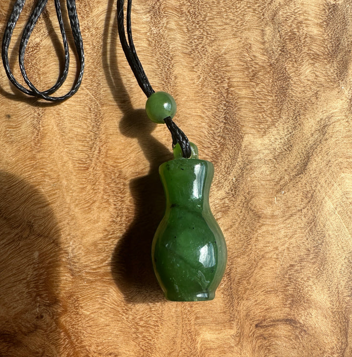 A green jade pendant shaped like a small bottle or vase, hanging from a black cord with a single green bead, placed on a textured wooden surface.
