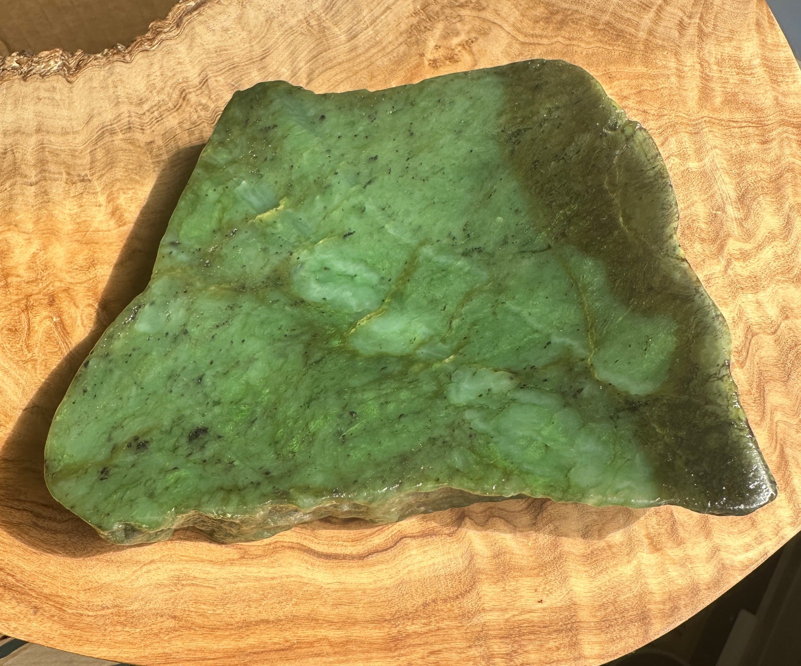 A polished green jade dish with natural marbling and dark veining, displayed on a curved wooden surface.