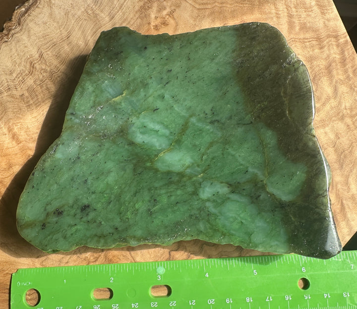 A polished green jade slab with natural marbling and dark veining, displayed on a wood surface with a green ruler at the bottom for scale.