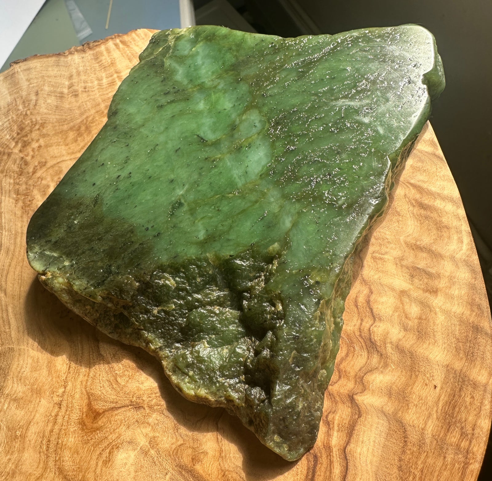 A polished green jade stone slab with natural marbling and rough edges, displayed on a curved wooden slice.