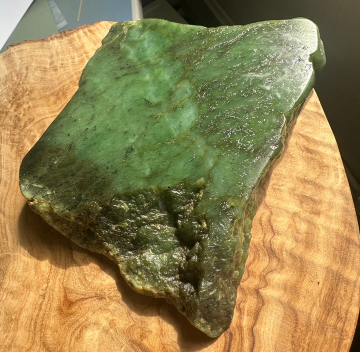 A polished green jade stone slab with natural marbling and rough edges, displayed on a curved wooden slice.