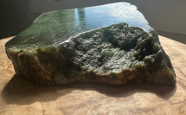 A raw polished green jade slab with a rough textured natural edge, resting on a wooden surface in warm natural light.