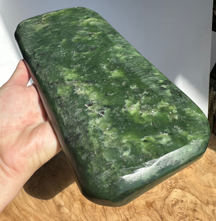 A hand holding a polished green stone block with natural marbled patterns, set against a background of a white wall and a wooden surface.
