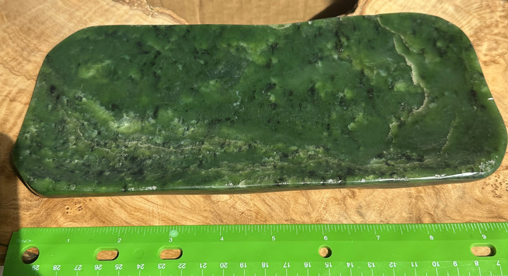 A green polished jade rectangular slab with rounded corners and natural marbling, shown on a wooden surface with a green ruler at the bottom for scale.