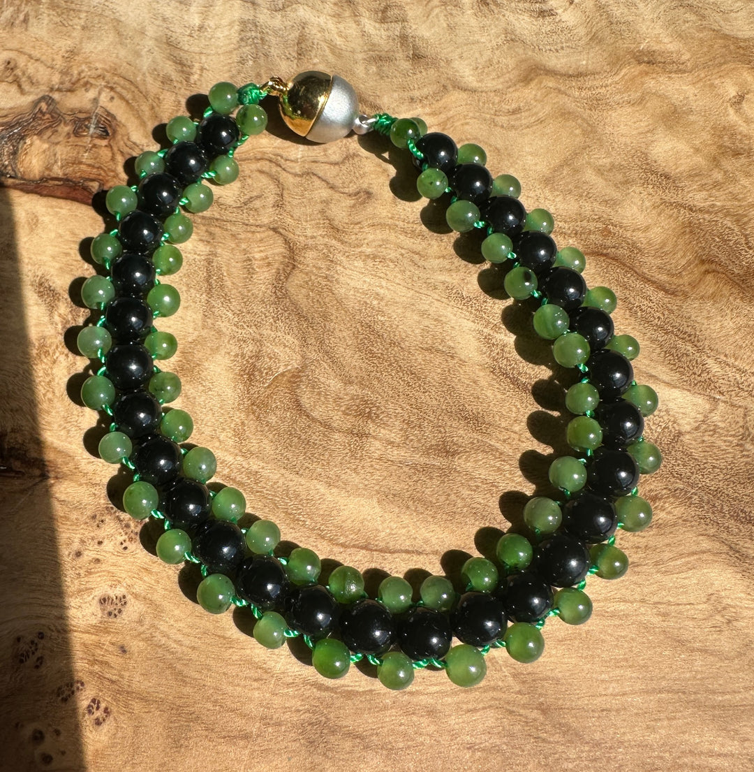 A beaded bracelet made of alternating green and black round beads, strung on a thin green cord and finished with a polished silver-toned bead clasp, displayed on a textured wood surface.