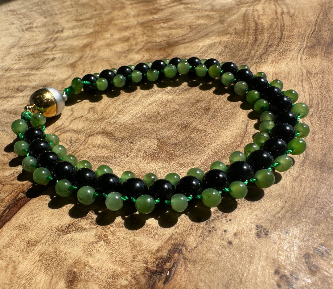 A beaded bracelet made of alternating black and green round beads, tied with a green cord and featuring a gold-toned accent bead, displayed on a textured wooden surface.
