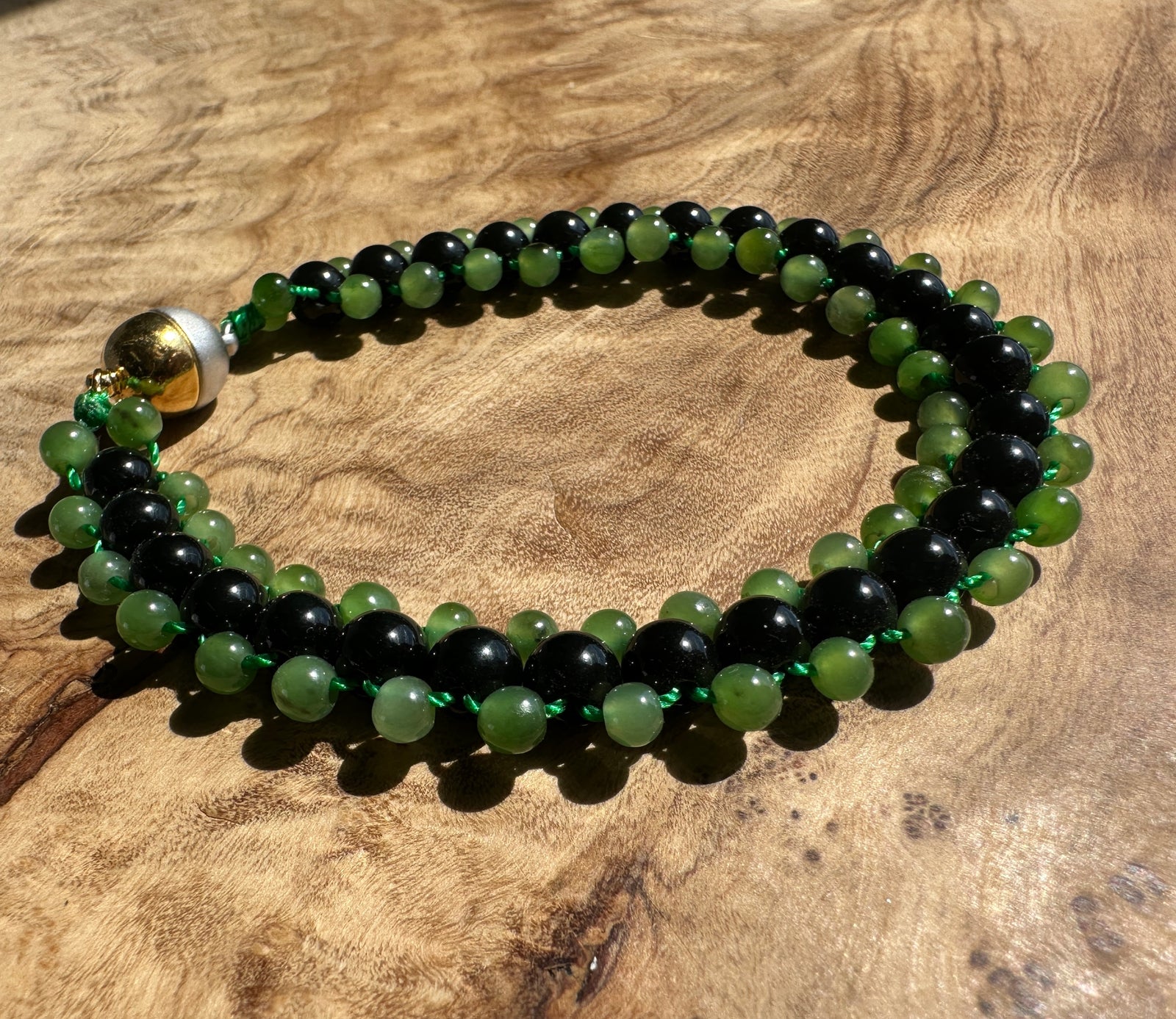 A beaded bracelet made of alternating black and green round beads, tied with a green cord and featuring a gold-toned accent bead, displayed on a textured wooden surface.