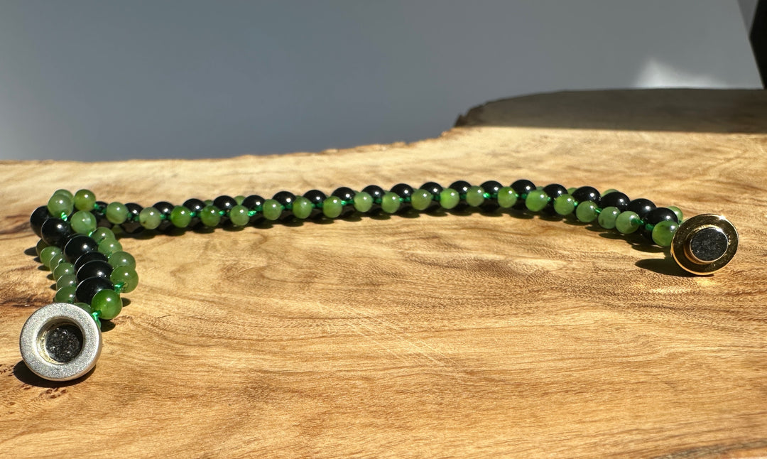 A beaded bracelet with alternating green and black round beads, featuring a silver-toned textured spacer bead on one end and a gold-toned spacer bead on the other, displayed on a rustic wood slice background.