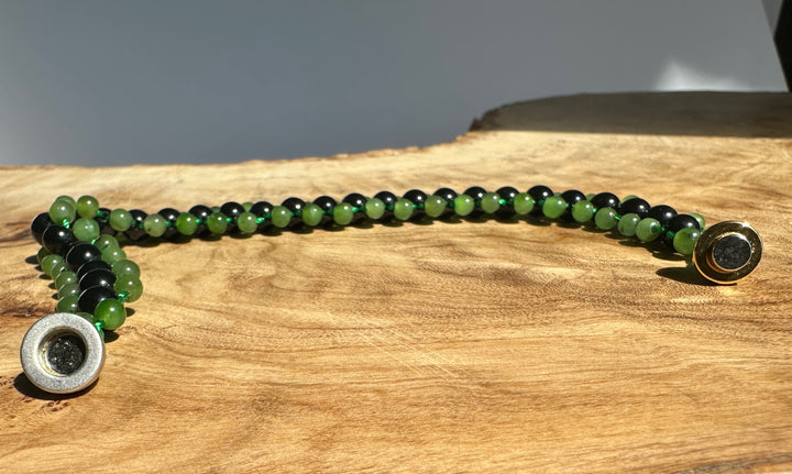 A beaded bracelet with alternating green and black round beads, featuring a silver-toned textured spacer bead on one end and a gold-toned spacer bead on the other, displayed on a rustic wood slice background.