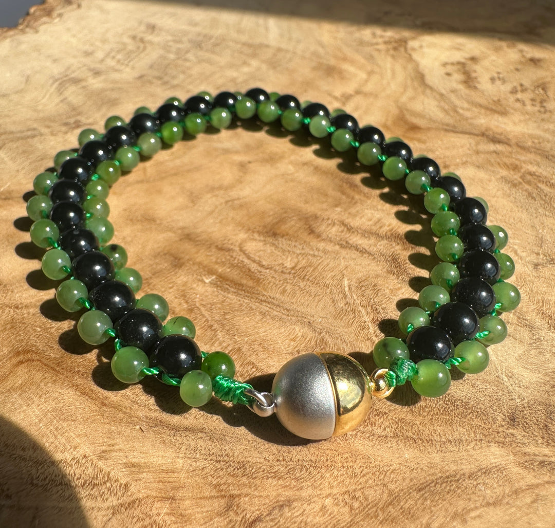 A beaded necklace with alternating green and black round beads, tied with green cord and featuring a large polished silver and gold bell centerpiece, displayed on a rustic wood slice background.