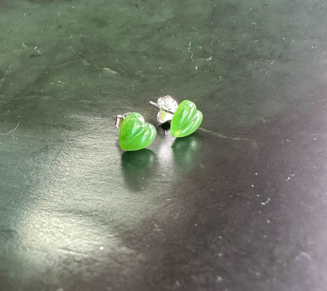 Two green jade heart stud earrings with silver posts on a dark surface