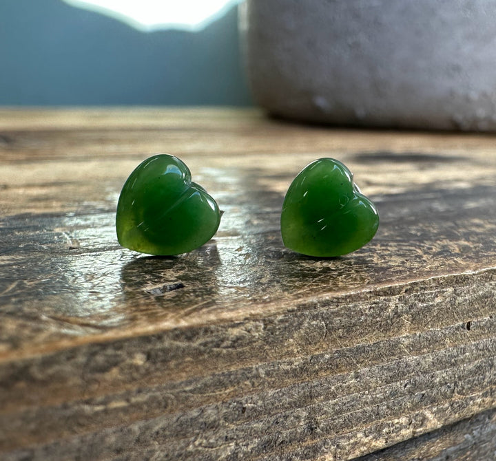 Two green heart shaped earrings with silver posts displayed on a weathered wooden surface