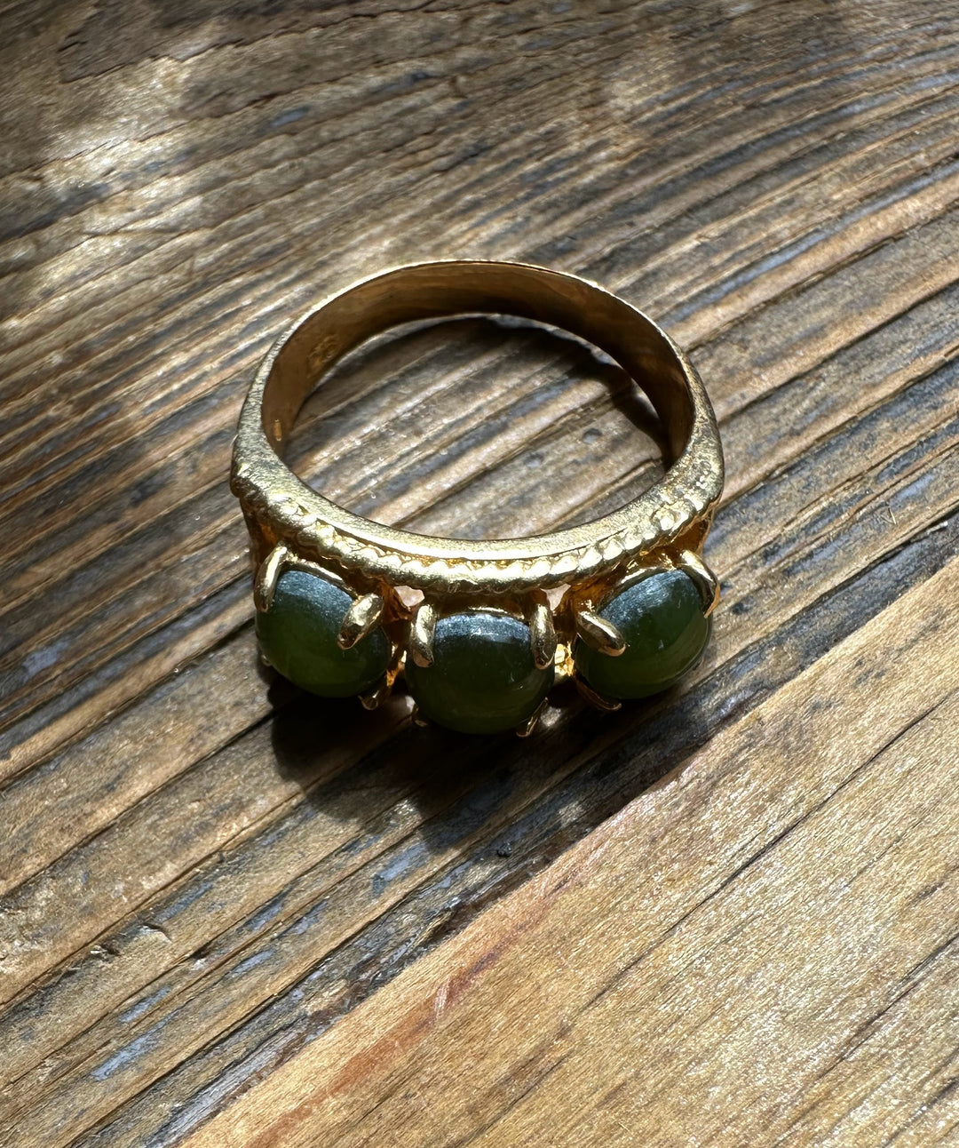 Gold ring featuring three green jade cabochons set in a row on a textured band on a wooden surface