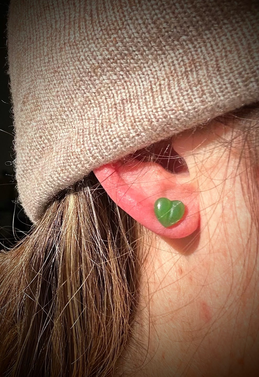 Green heart shaped stud earring worn on the earlobe under a beige hat