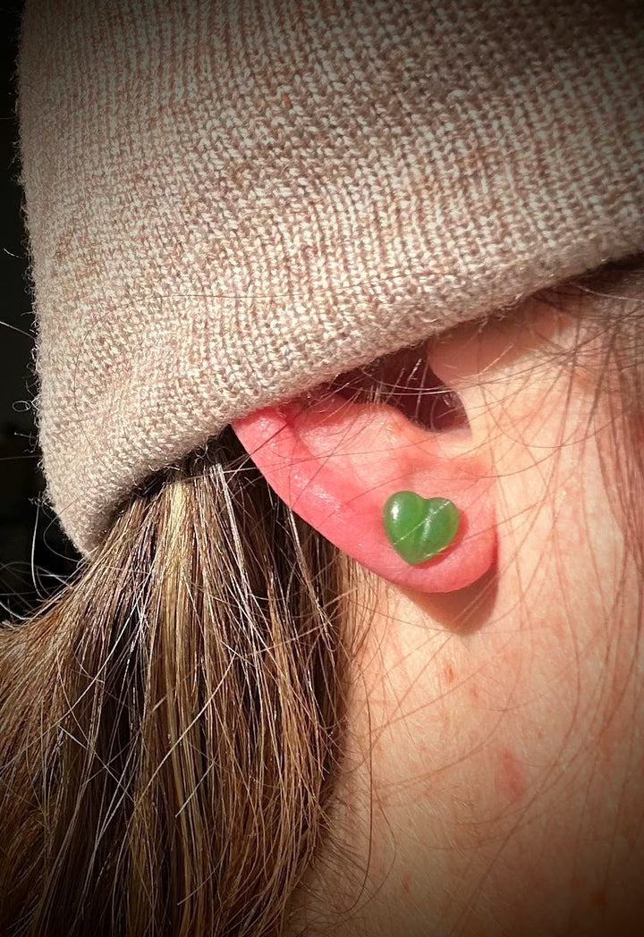 Green heart shaped stud earring worn on the earlobe under a beige hat