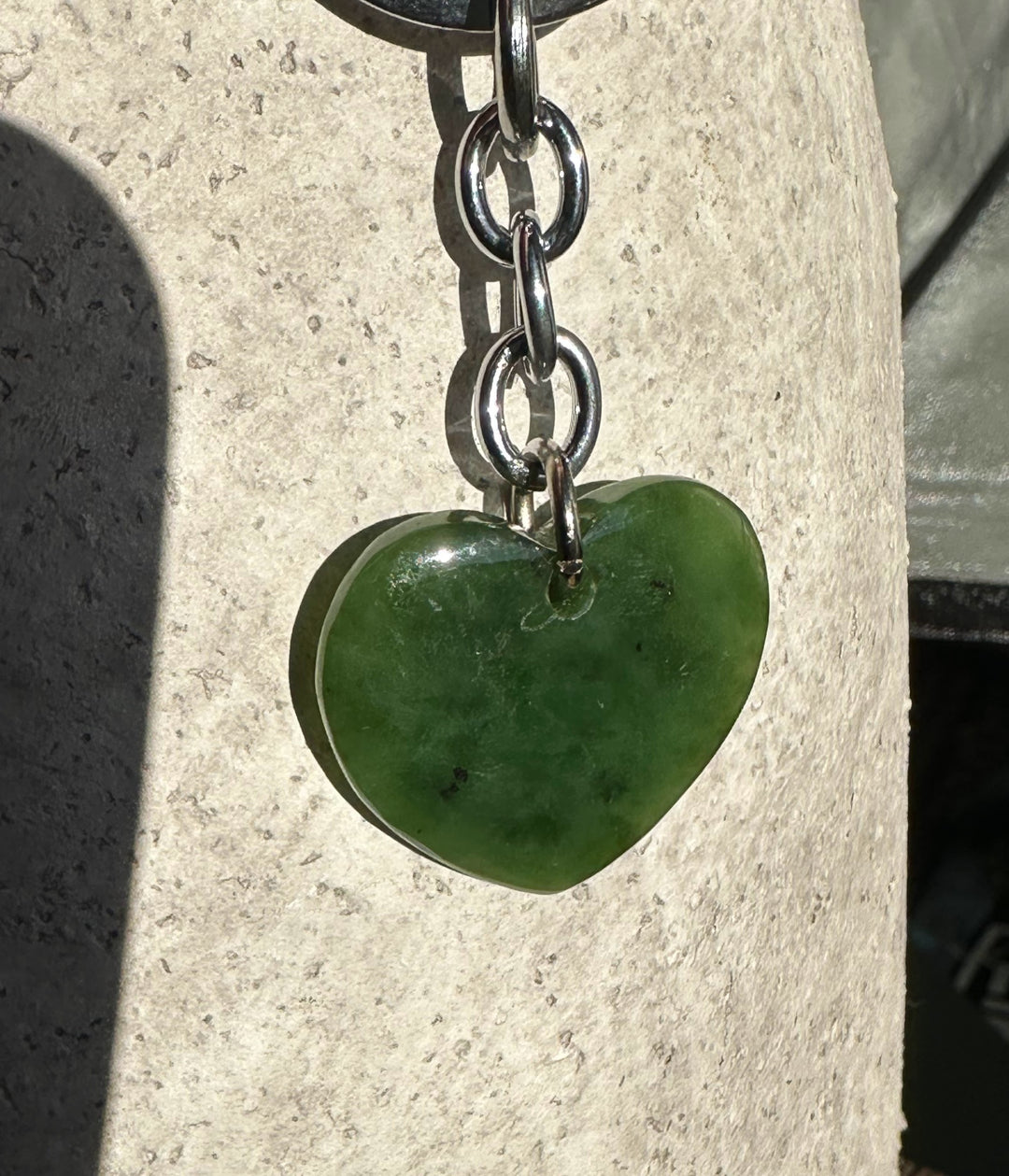 Close up of a green heart pendant on a metal chain with a large silver keyring above, resting on a light stone surface.