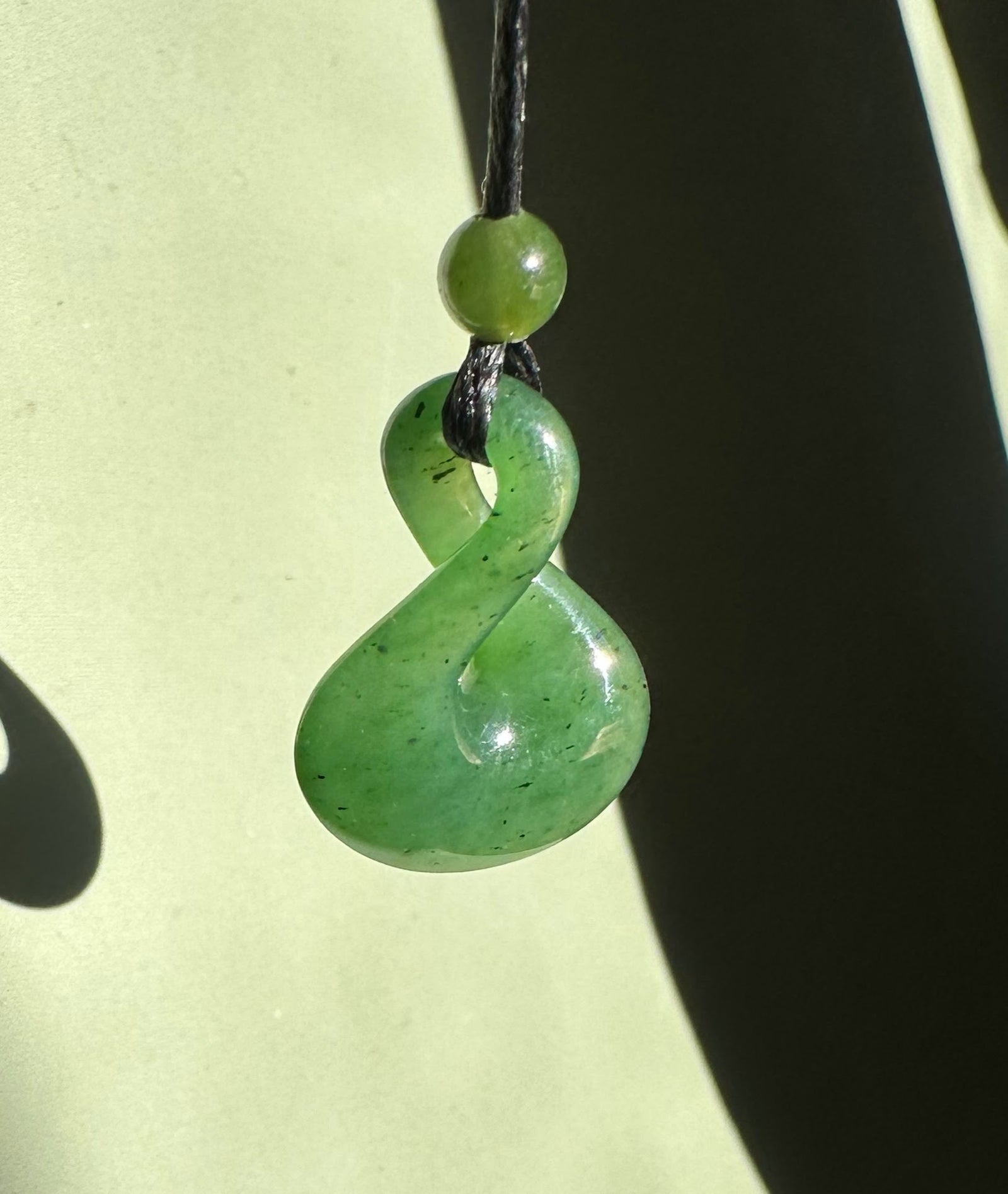 Green jade infinity twist pendant suspended on a black cord with a small round bead above