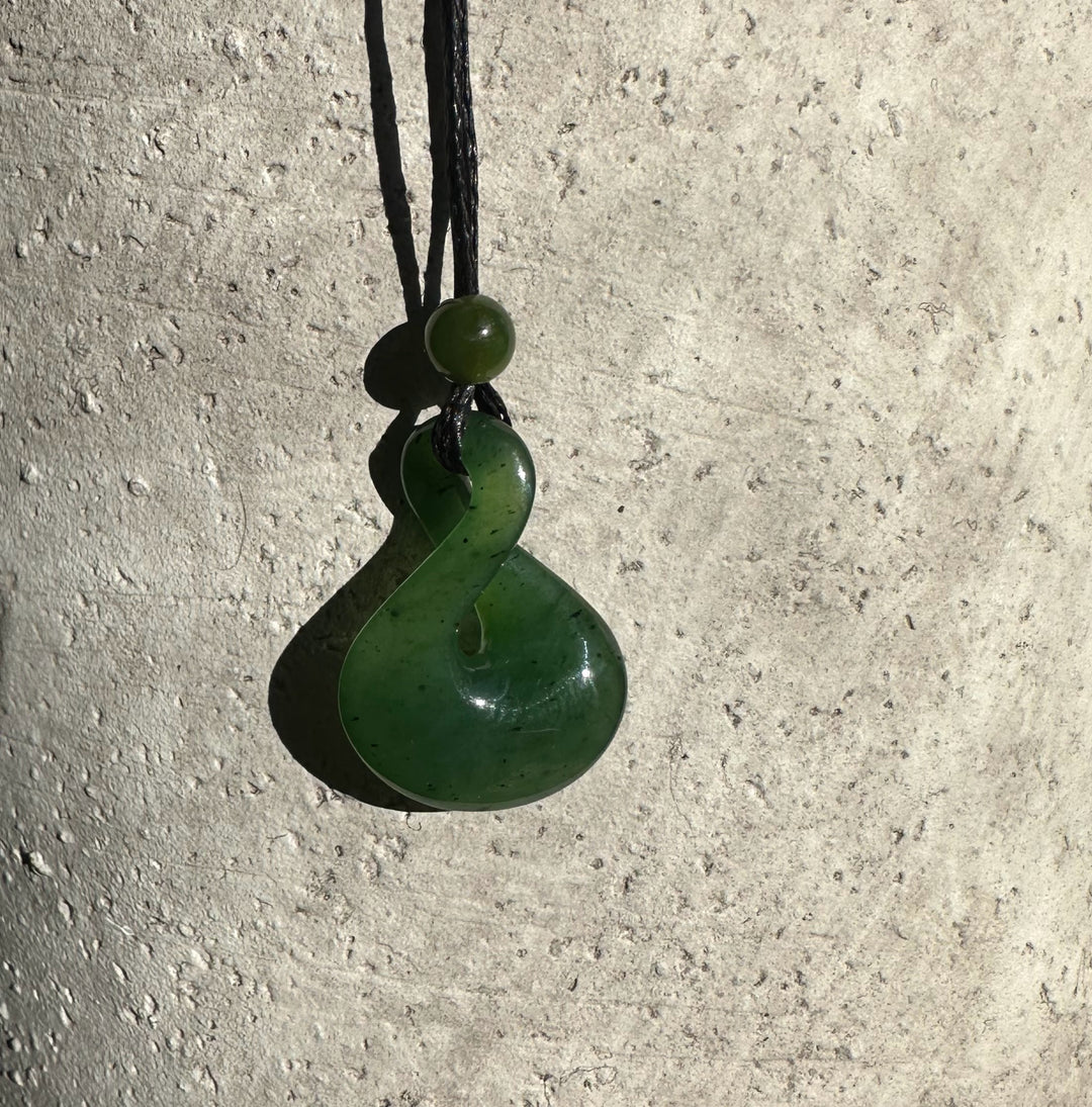 Green jade infinity twist pendant on black cord against a light textured wall.