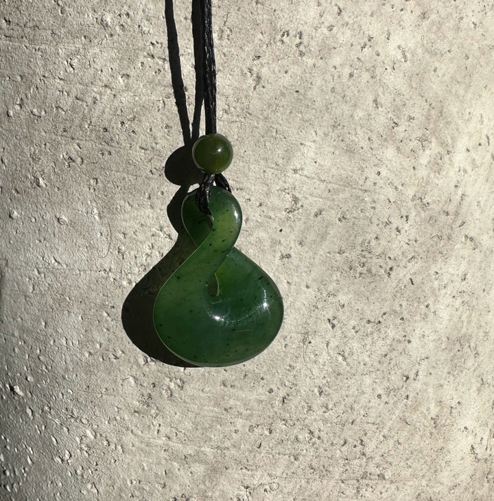Green jade infinity twist pendant on black cord against a light textured wall.