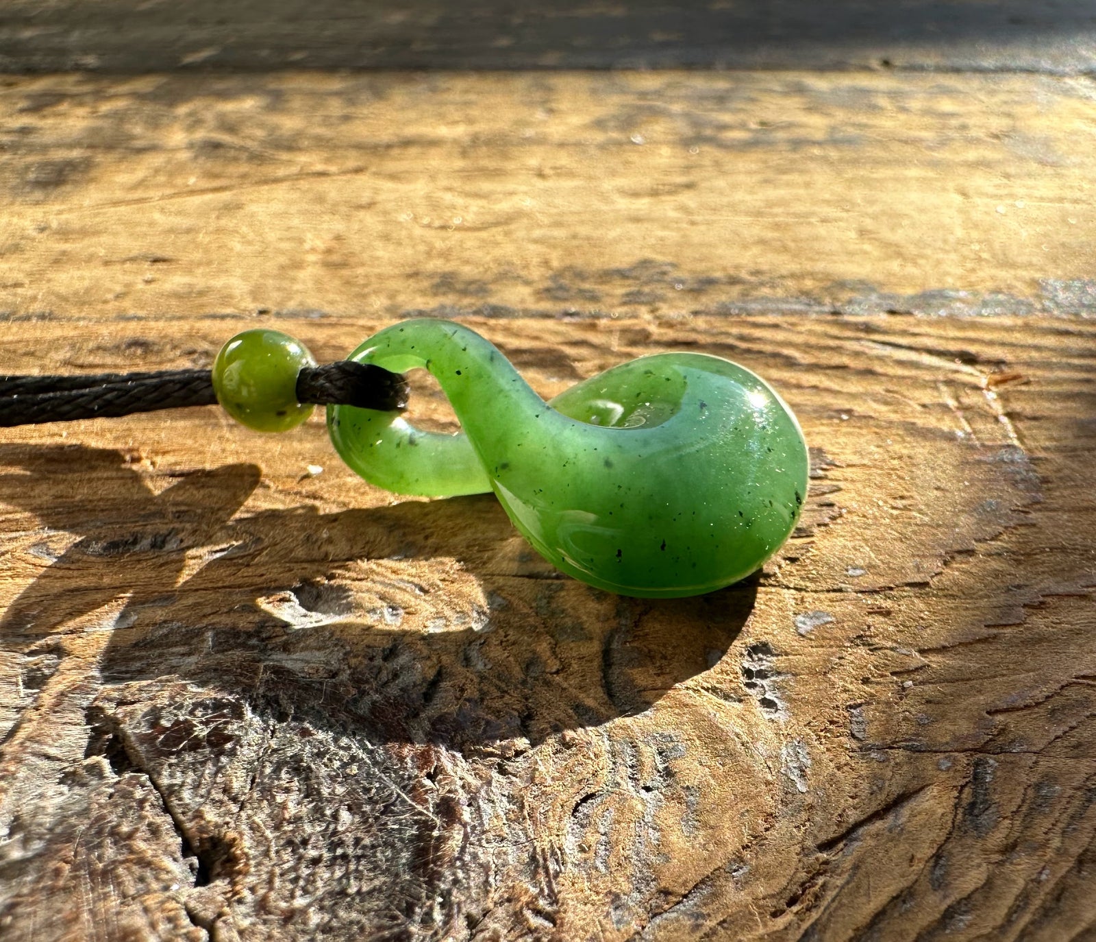 Green jade infinity twist pendant on rustic wood surface bathed in sunlight
