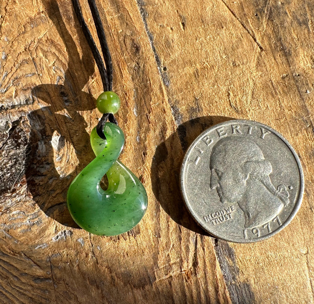 Green jade infinity twist pendant with bead on black cord placed next to a coin for scale.