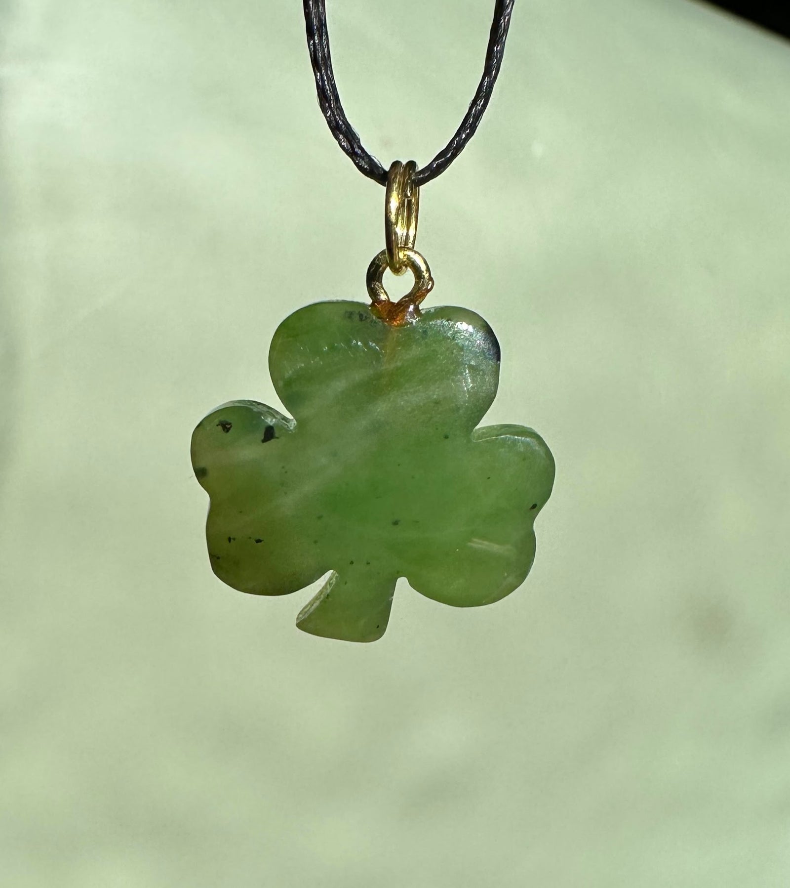 Close up of a green jade three leaf clover pendant hanging from a black cord with a gold bail