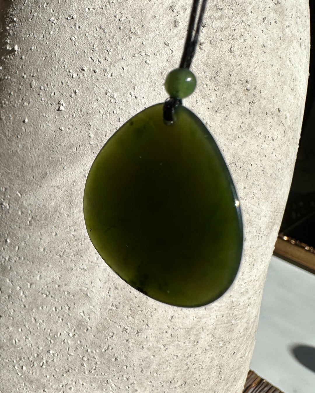 Close-up of dark green jade teardrop pendant on a black cord with a green bead against a textured light background