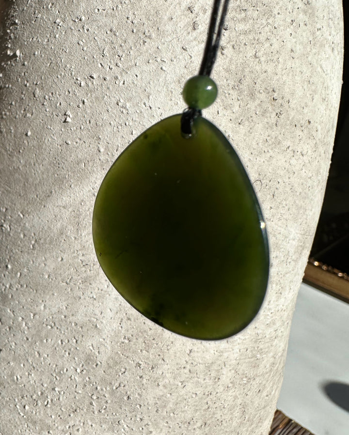 Close-up of dark green jade teardrop pendant on a black cord with a green bead against a textured light background