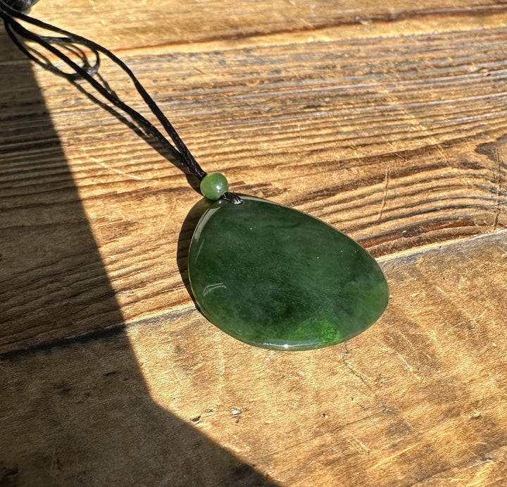 Dark green jade pendant with a small green bead on a black cord resting on a sunlit wooden surface