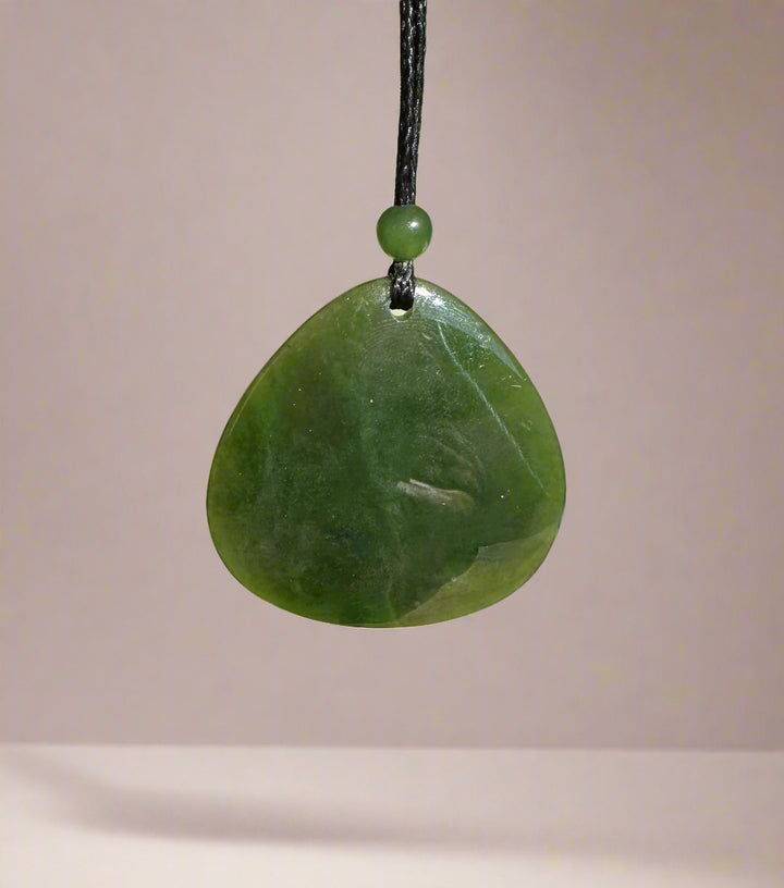 Dark green jade pendant suspended on a black cord with a small green bead above, against a pale pink background