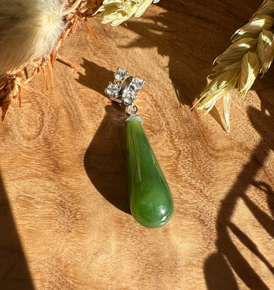 Green nephrite jade teardrop pendant with a silver cap and crystal accents on a wood surface.