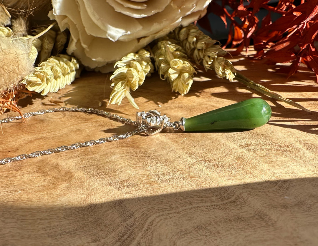 Green nephrite jade teardrop pendant with silver setting and chain resting on a wooden surface.