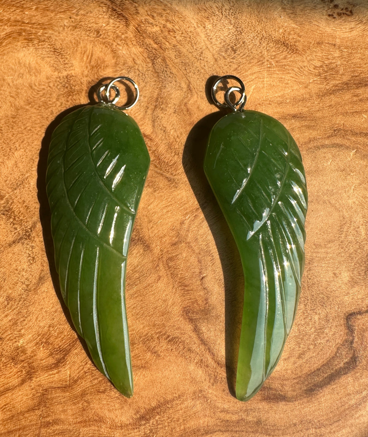 Two green jade angel pendant with silver-colored hanging loops, displayed on a textured wooden surface.