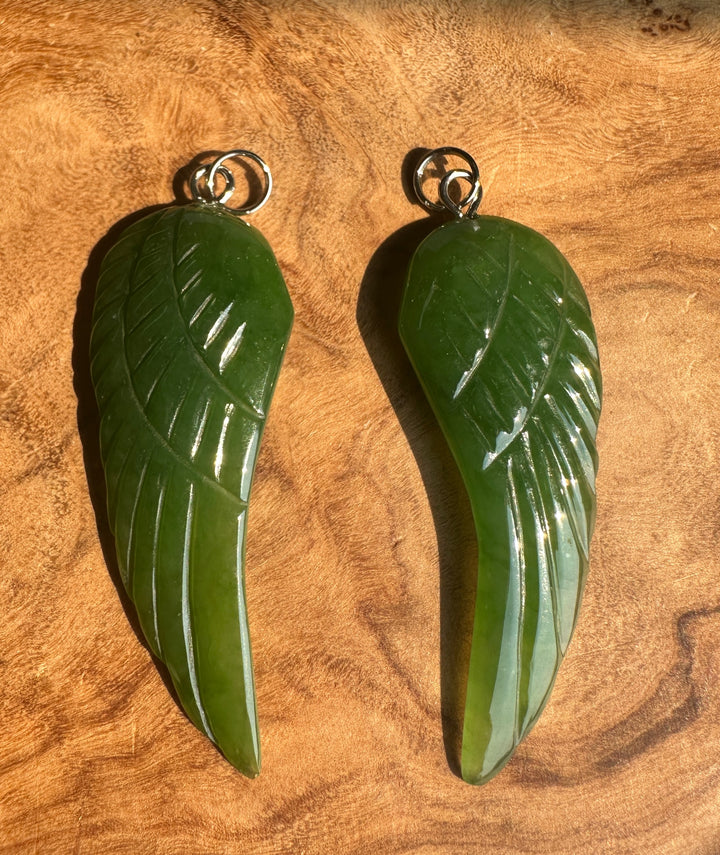 Two green jade angel pendant with silver-colored hanging loops, displayed on a textured wooden surface.
