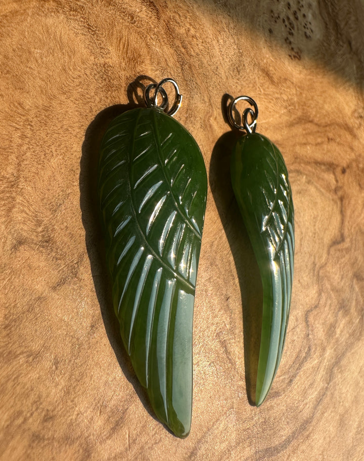 Two green carved wing pendants with silver jump rings, displayed on a textured brown wood background.