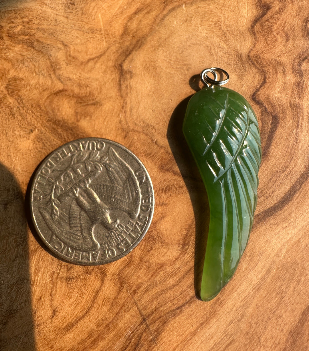 A carved green jade pendant shaped like a wing with a silver ring, displayed next to a silver coin on a textured olive wood background.
