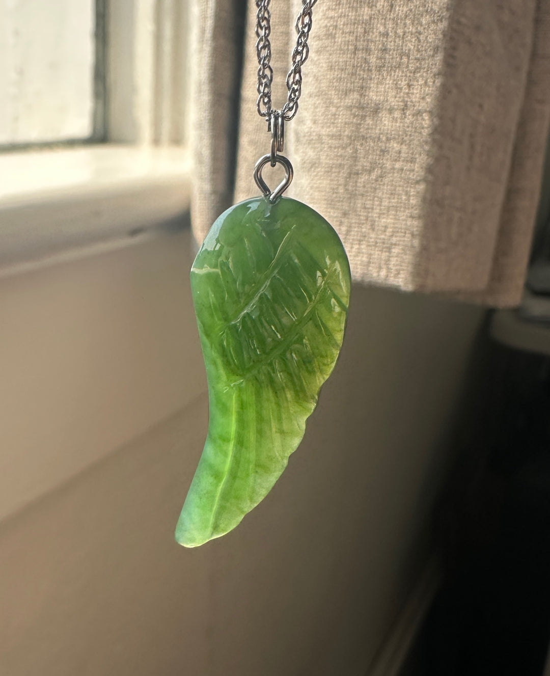 A green carved wing-shaped pendant necklace hanging from a silver chain in front of a window with a beige curtain.