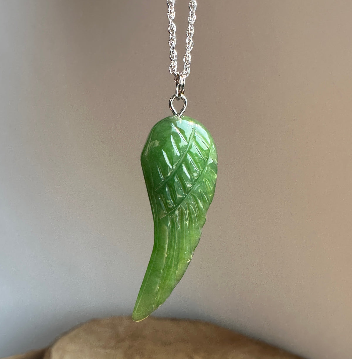 A green carved stone wing pendant necklace on a silver chain, shown against a neutral gray background.