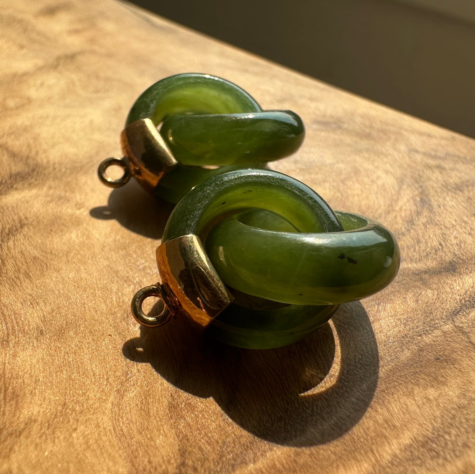 Two green jade  charms with gold-toned metal loops, resting on a wooden surface under direct sunlight.