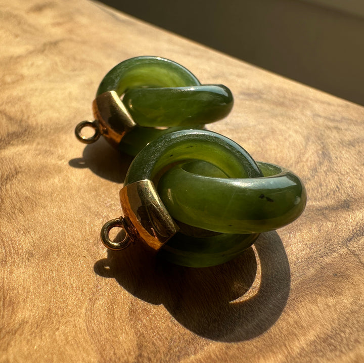 Two green jade  charms with gold-toned metal loops, resting on a wooden surface under direct sunlight.