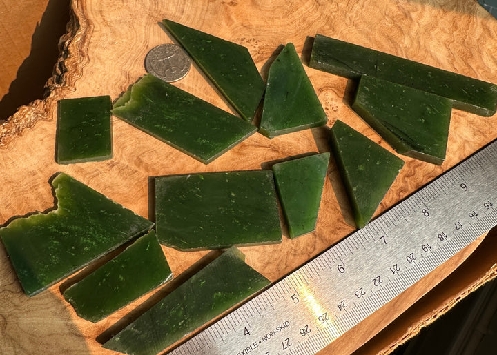 Several dark green translucent rectangular and triangular jade scattered on a wood slice surface, with a coin and metal ruler for scale.