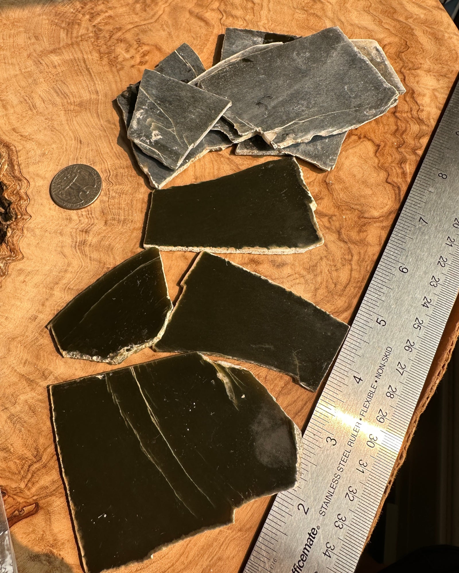 Several dark jade slabs natural edges are arranged on a wood slab, with a coin and a metal ruler nearby for scale.