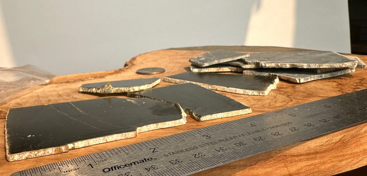 Several dark rectangular jade with rough light-colored edges are arranged on a wooden surface next to a metal ruler for scale.