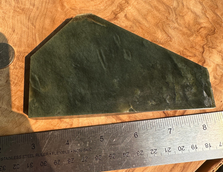 A dark green stone with natural striations, shown on a wood-grain surface next to a stainless steel ruler for scale.