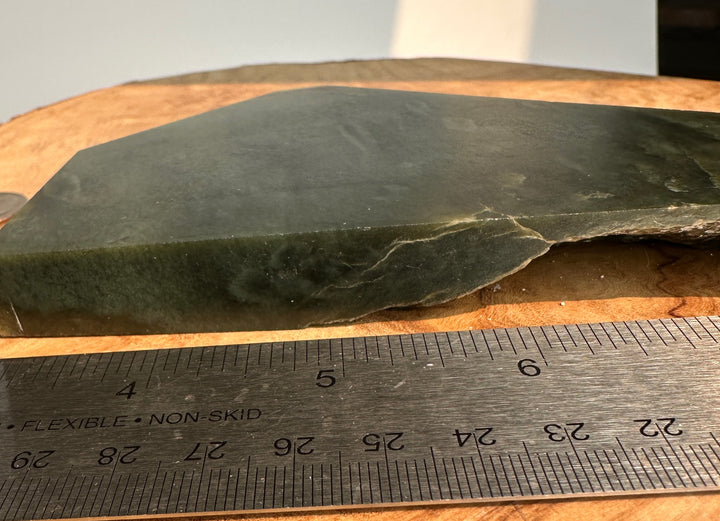 A dark green stone  shown on a wooden surface with a metal ruler in front for scale.