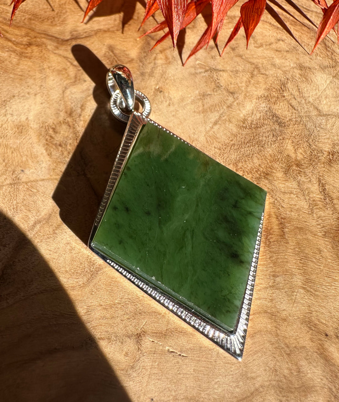 Green jade pendant set in a square silver bezel with a beaded edge and bail, on a wooden surface with red leaves.