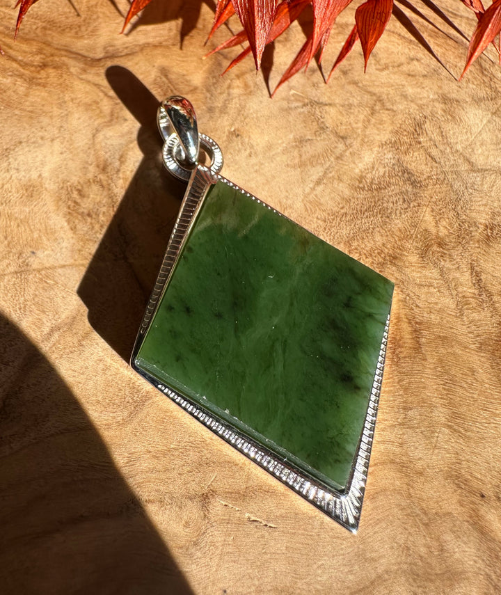 Green jade pendant set in a square silver bezel with a beaded edge and bail, on a wooden surface with red leaves.