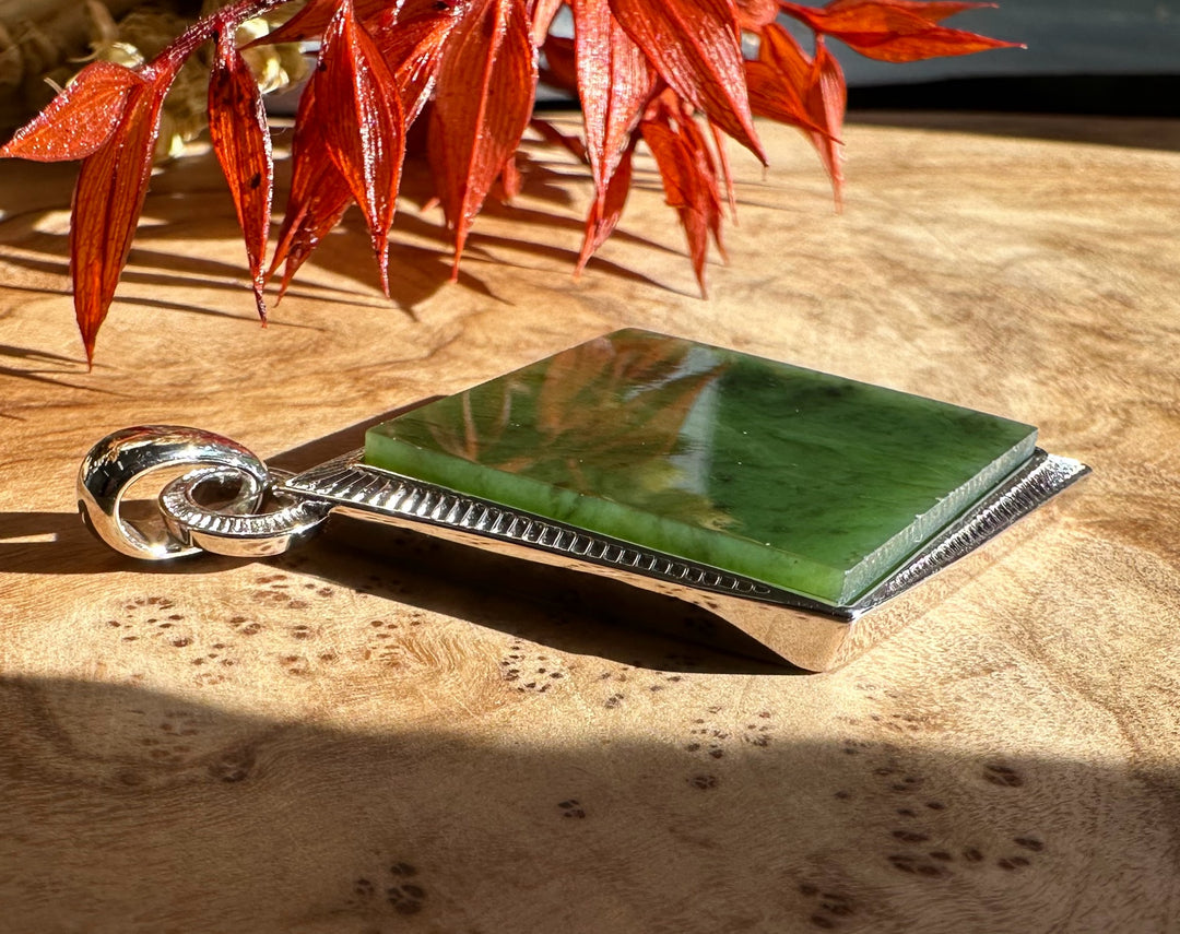 Green jade square pendant with a silver border resting on a wood surface with red leaves above