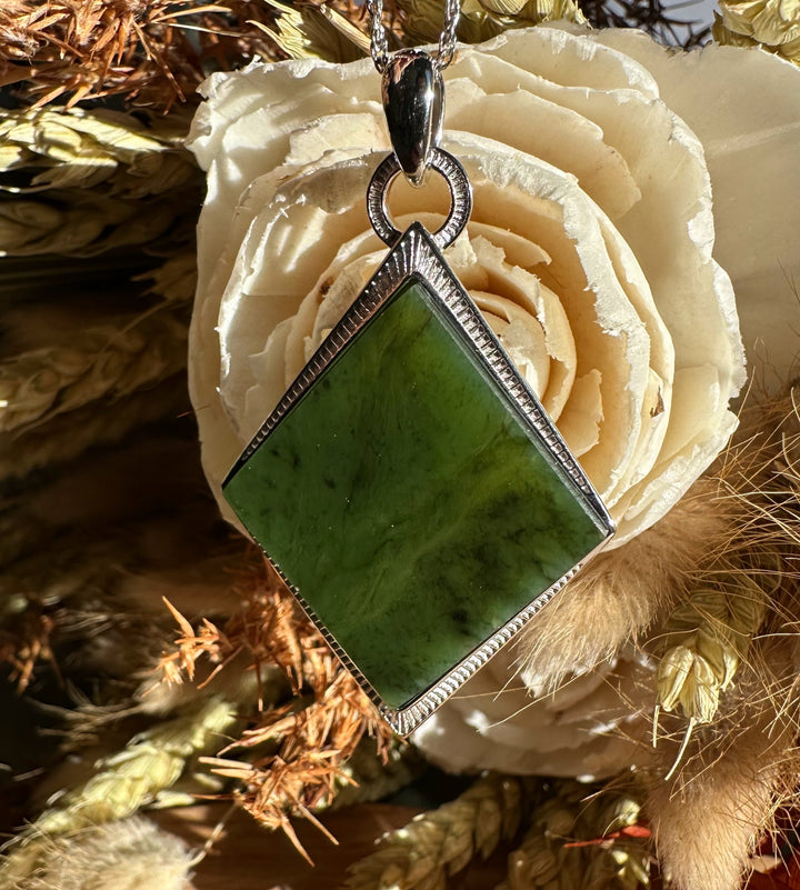 Diamond shaped green jade pendant with a textured silver bezel on a chain against a cream rose background