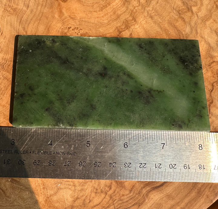 A rectangular green jade slab with natural marbling, shown on a wooden surface with a metal ruler underneath for scale.